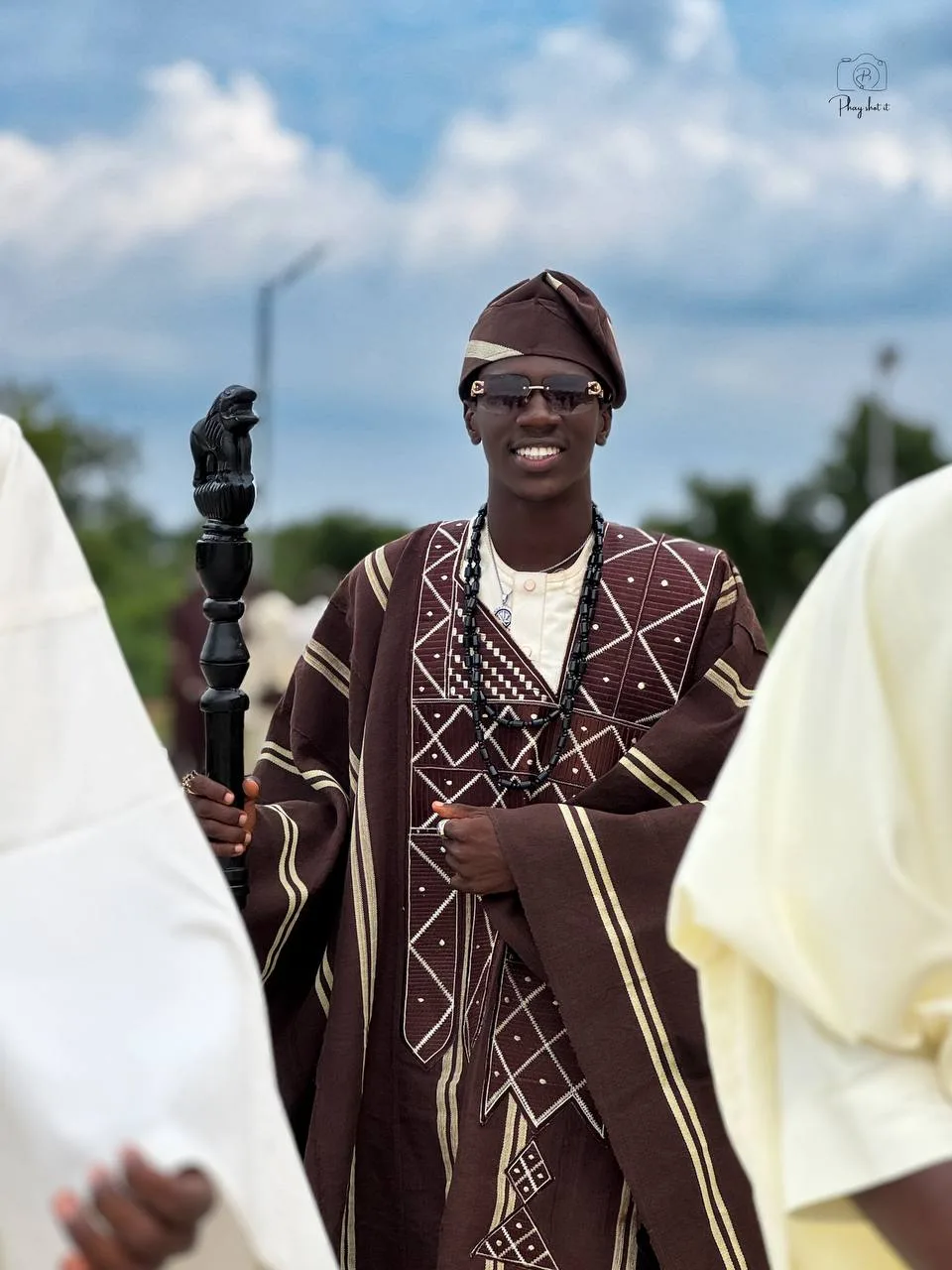 Young man in traditional brown Yoruba agbada with carved staff, outdoor portrait in Ilorin.