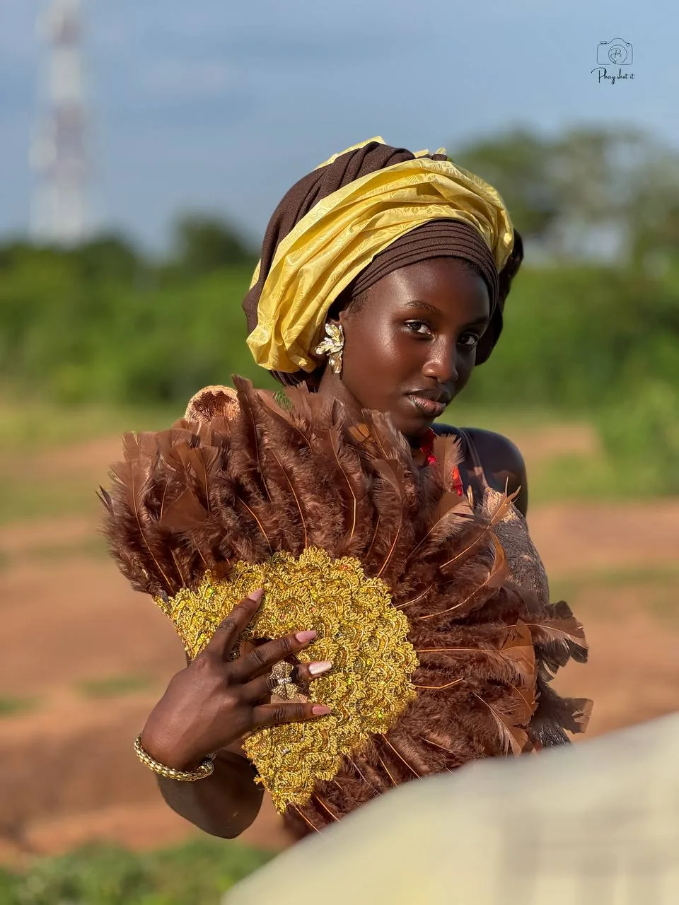 Woman in gold gele headwrap and brown feathered attire with gold beaded fan, outdoor portrait.