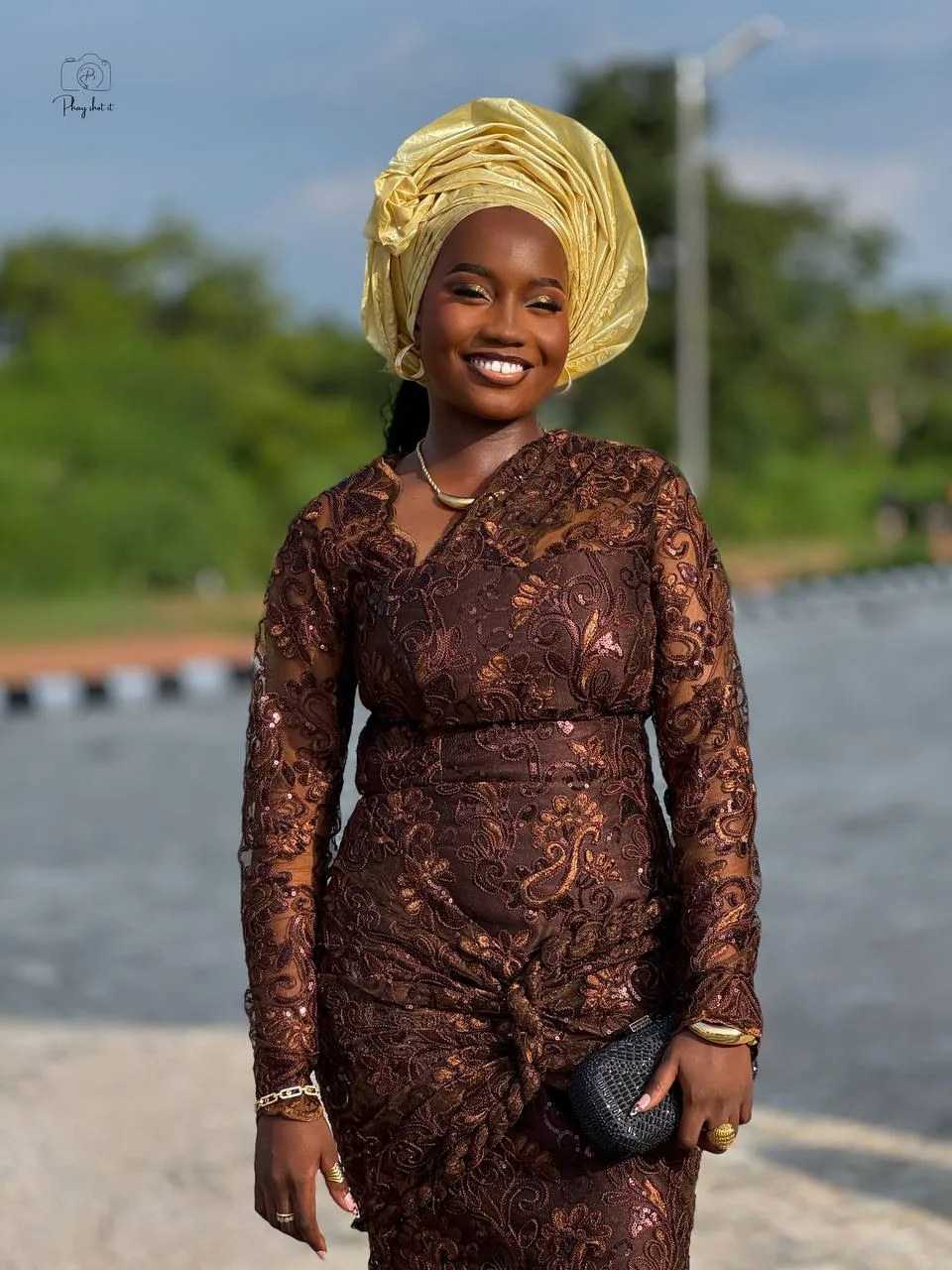 Smiling woman in embroidered brown lace dress with gold gele headwrap, standing outdoors.