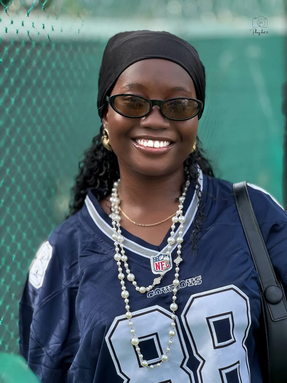 Smiling woman in navy Dallas Cowboys jersey with pearl necklaces against green fence.