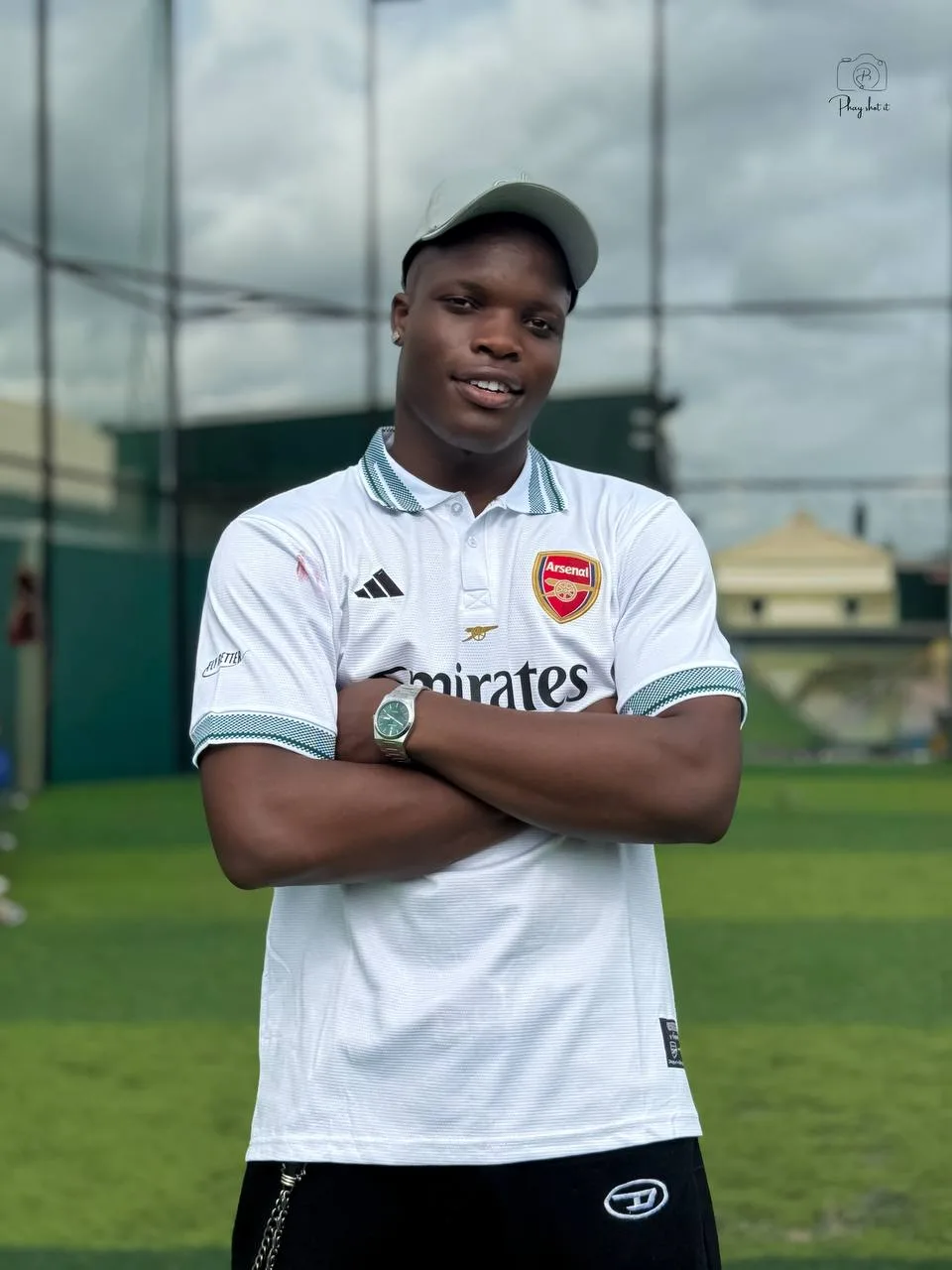 Man in white Arsenal jersey standing with arms crossed on turf pitch, wearing cap and watch.