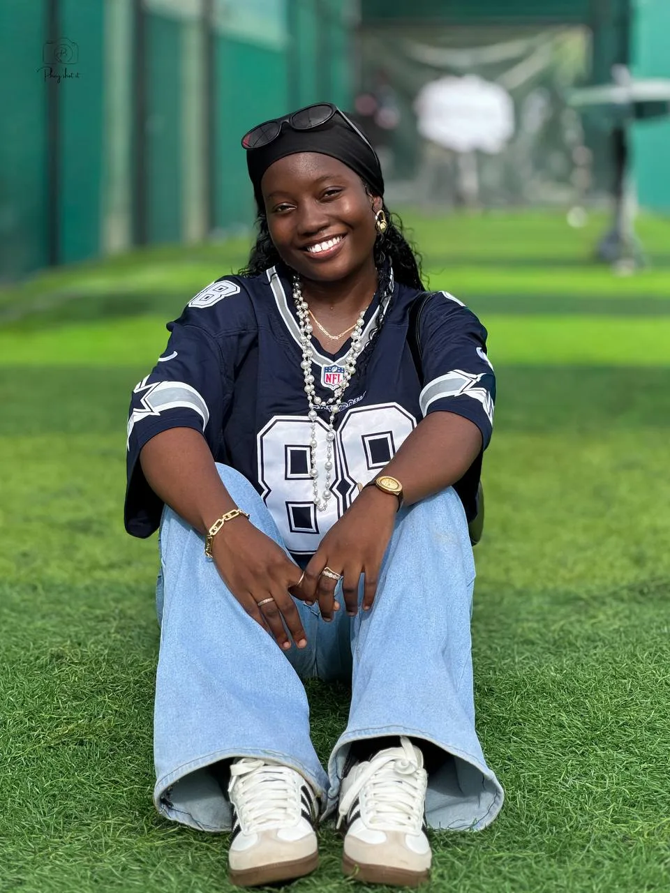Woman in Dallas Cowboys jersey seated on grass with jeans and white sneakers, smiling.