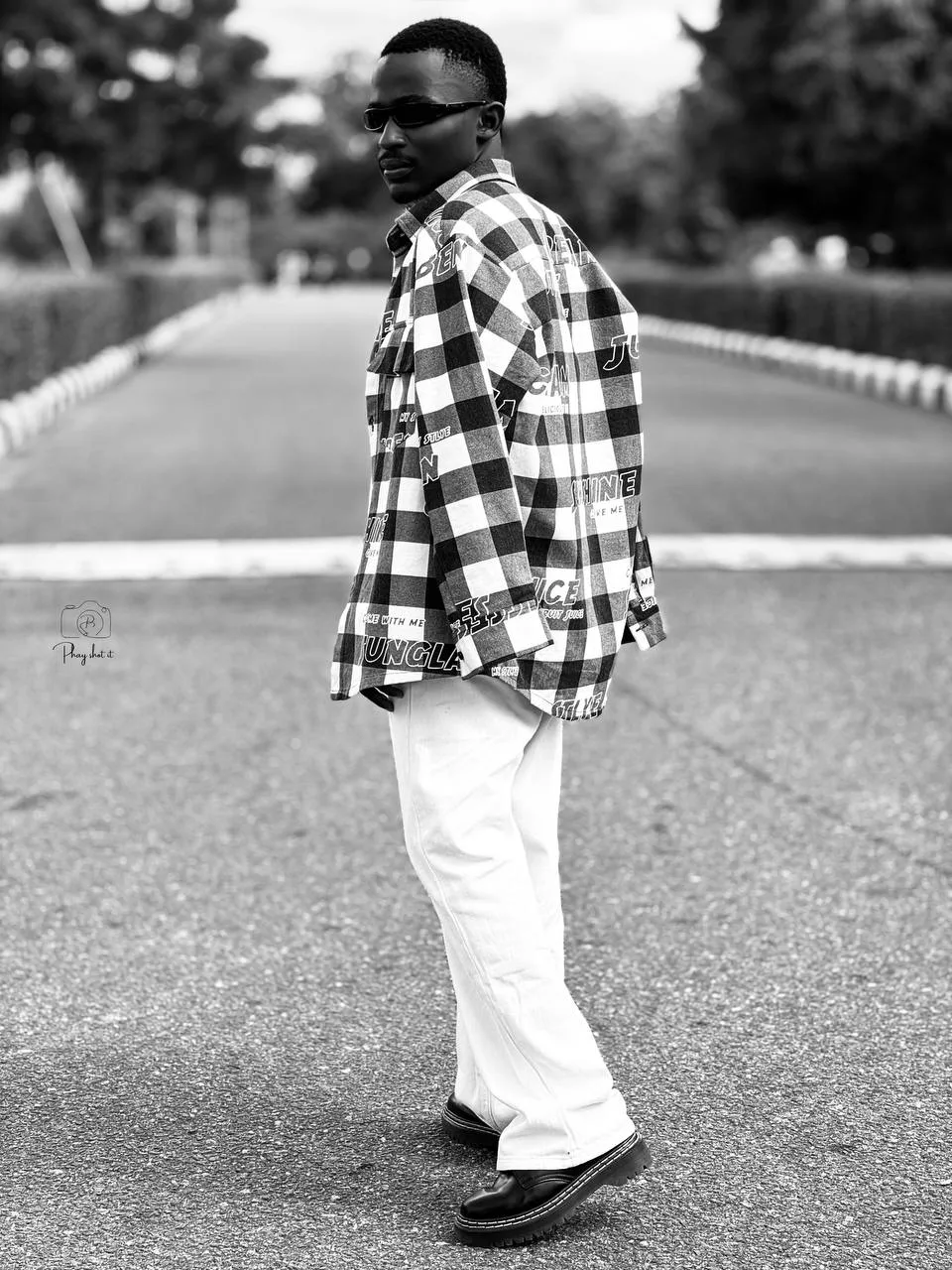 Man in oversized check shirt and white trousers walking on a long road, black and white portrait.