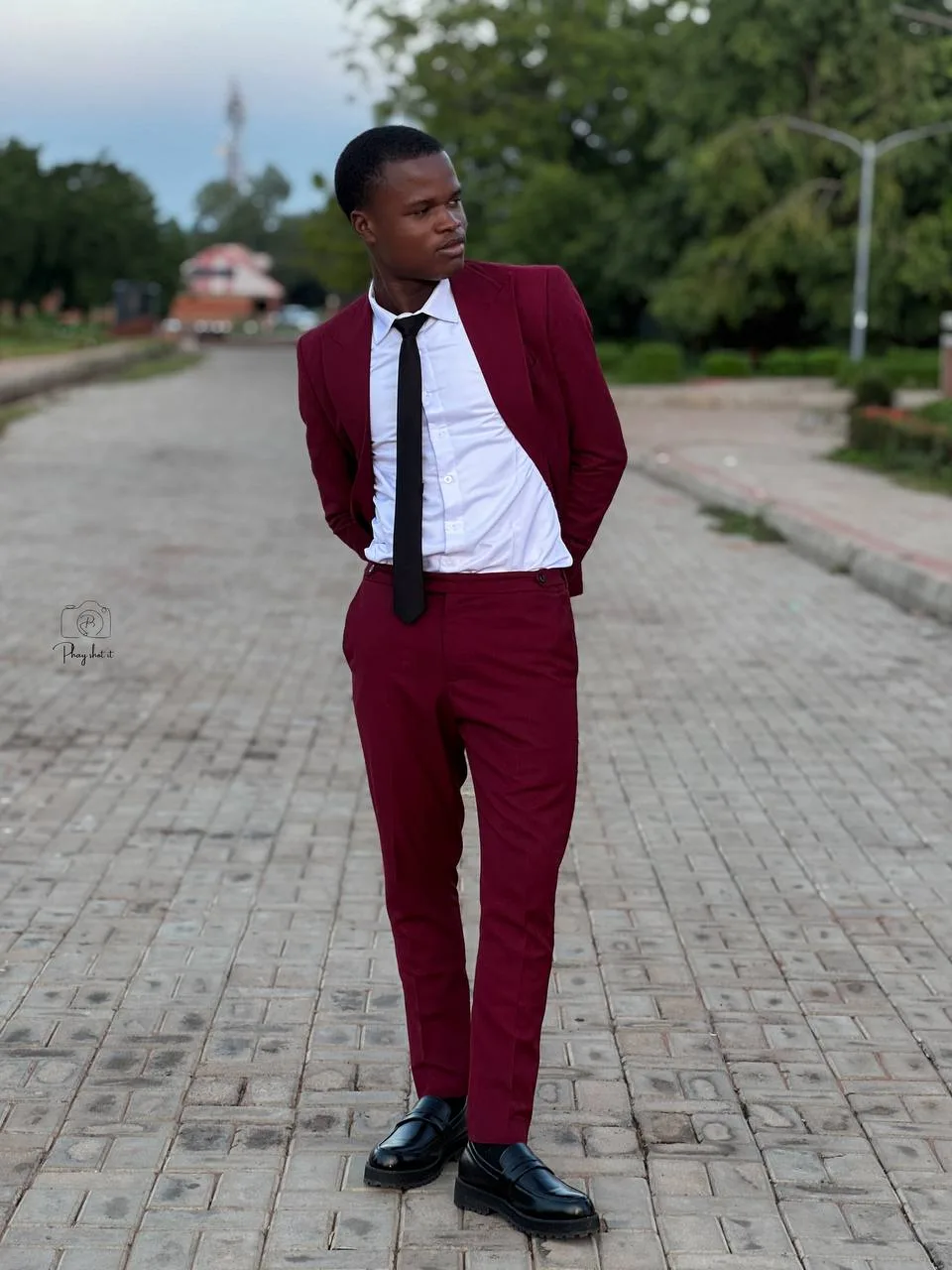 Man in burgundy suit and white shirt with black tie, standing on brick pathway looking away.
