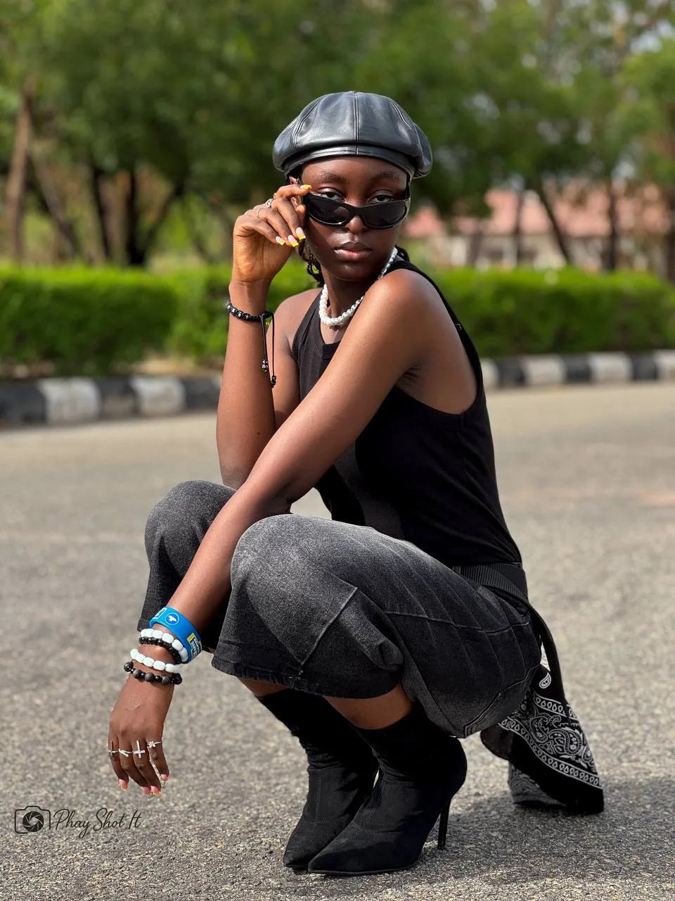Woman crouching on road in all-black outfit with leather beret and sunglasses, street style.