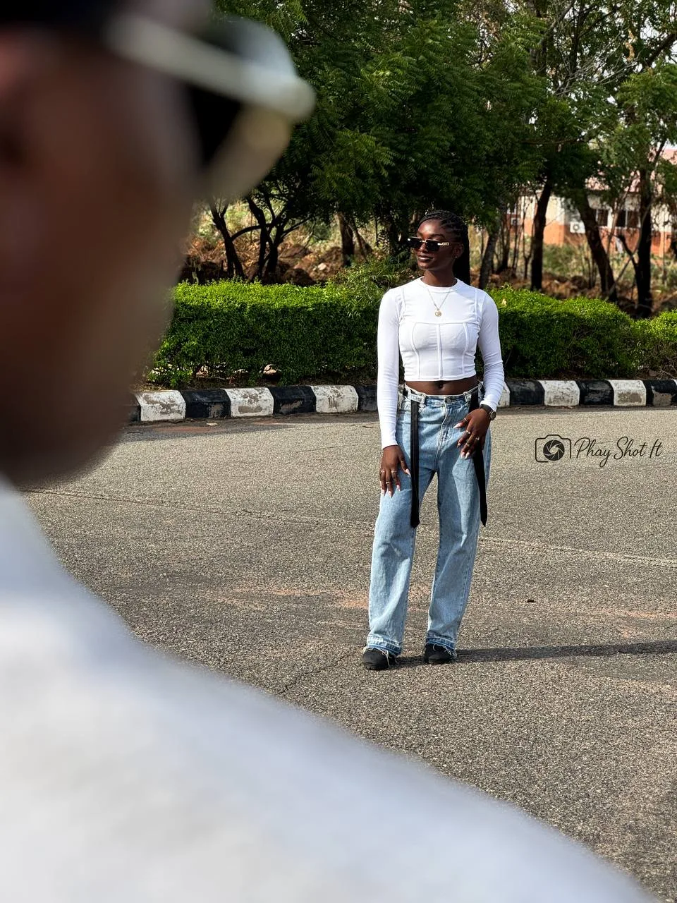 Woman standing in white crop top and blue jeans on paved road, shot through foreground blur.
